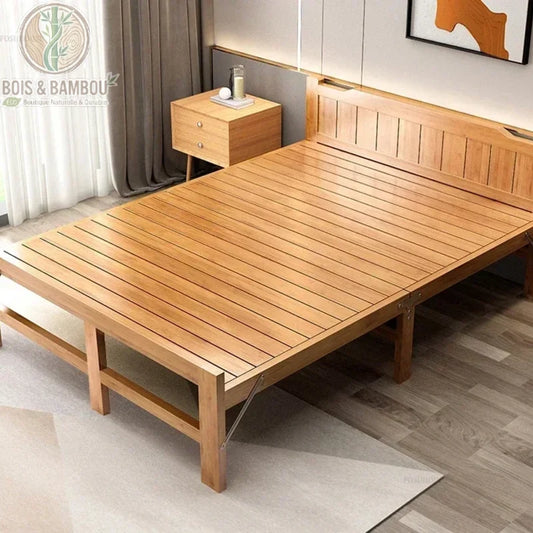 Lit Pliant en Bambou Massif 150x197. Large lit double pliant en bambou massif verni avec sommier à lattes, présenté dans une chambre moderne à côté d'une table de chevet assortie sur un tapis clair.