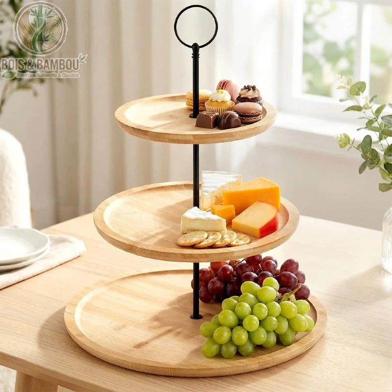 Présentoir en Bambou à 3 Étages. Un serviteur muet avec plateaux en bambou et tige noire, garni de pâtisseries, fromages et raisins, posé sur une table en bois claire.