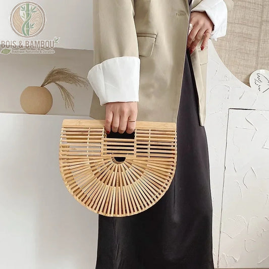 Une femme portant une veste beige et une robe noire tient un sac à main demi-lune en bambou ajouré, illustrant un style élégant et naturel.