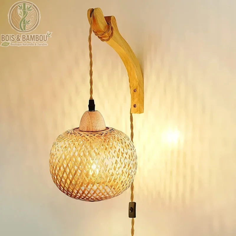 Applique murale en bambou tressé arborant une forme de lanterne sphérique suspendue par un cordon torsadé. Fixée sur une potence courbée en bois clair, elle projette des ombres géométriques élégantes sur le mur grâce à son tressage artisanal ajouré.