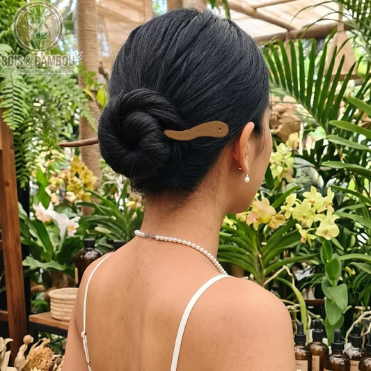 Bâton à cheveux en bois de style chinois. Une femme de dos, coiffée d'un chignon serré maintenu par un bâton à cheveux en bois clair scuplté. Elle porte un collier de perles et des boucles d'oreilles pendantes assorties, devant un arrière-plan luxuriant de plantes tropicales et d'orchidées jaunes.