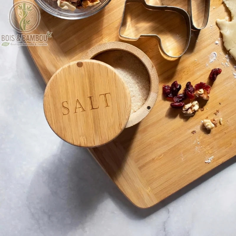 Boîte à sel en bambou avec couvercle pivotant gravé, posée sur une planche à découper. Ce récipient naturel et compact permet de conserver le sel à portée de main tout en ajoutant une note boisée et épurée à votre plan de travail.