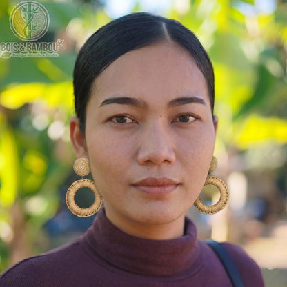 Boucles d'oreilles bohèmes en rotin et bois circulaires portées par une femme en gros plan, avec un arrière-plan naturel flou.