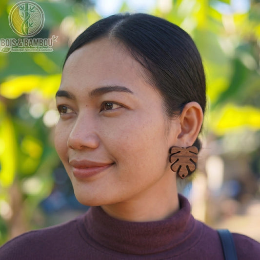 Boucles d'oreilles en bois naturel en forme de feuille tropicale portées par une femme de profil, avec un arrière-plan naturel flou.