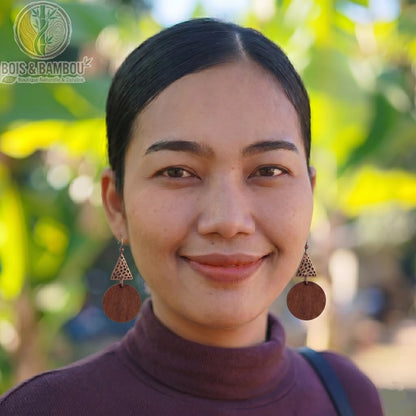 Boucles d'oreilles ethniques en bois naturel portées par une femme en gros plan, avec des détails géométriques et un arrière-plan naturel flou.