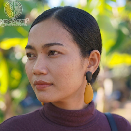 Boucles d'oreilles géométriques en bois noir et jaune portées par une femme de profil, avec une forme de goutte allongée et un arrière-plan naturel flou.