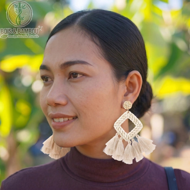 Boucles d'oreilles pendantes en bambou et rotin tressés avec pompons, portées par une femme de profil sur un fond naturel ensoleillé et verdoyant.