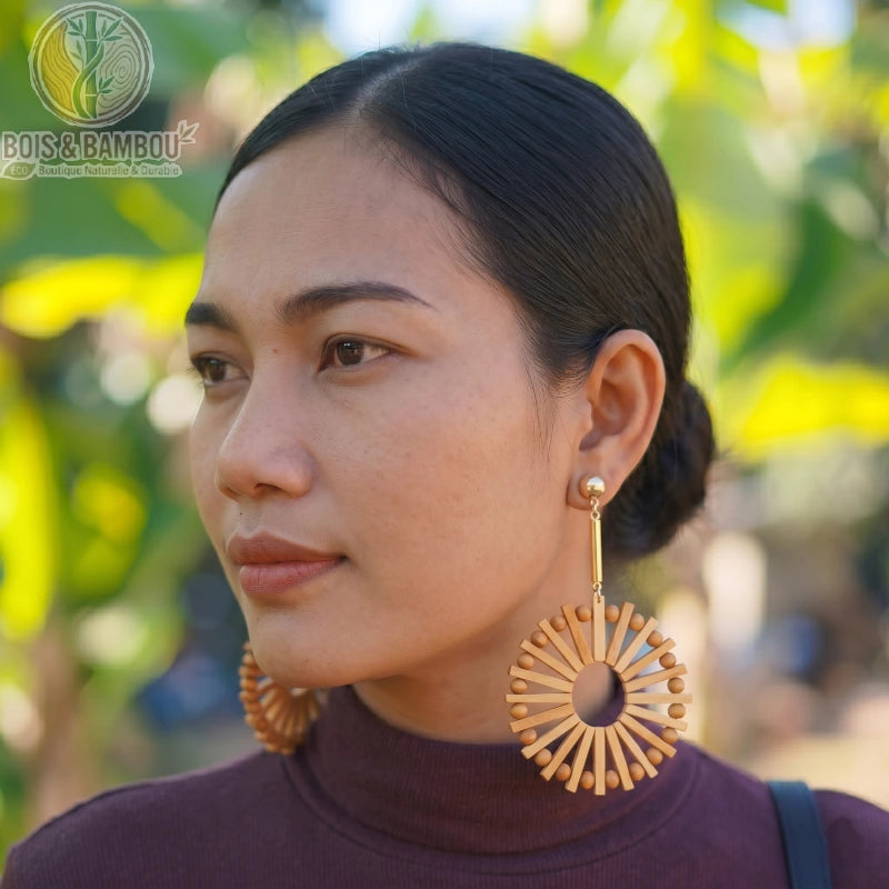 Boucles d'oreilles pendantes en rotin et bambou de forme circulaire portées par une femme de profil, avec un arrière-plan naturel flou.
