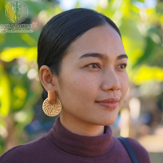 Boucles d'oreilles pendantes en rotin et bois de forme ronde portées par une femme de profil, avec un design tressé naturel et un arrière-plan flou.