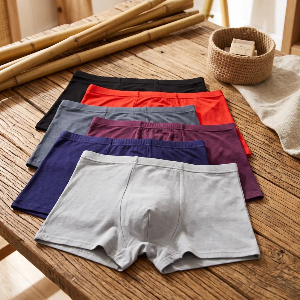 Six caleçons boxeurs pour hommes de différentes couleurs (gris clair, bleu marine, bordeaux, gris foncé, rouge et noir) disposée sur une table en bois rustique.