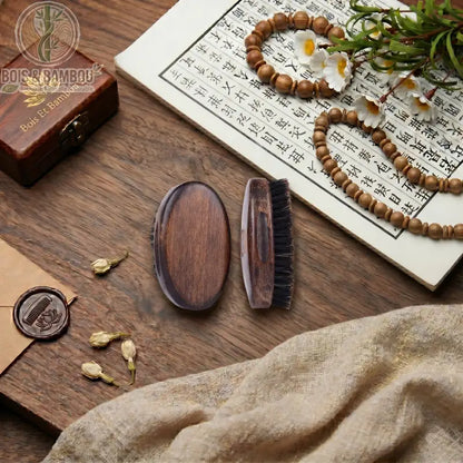 Brosse à barbe en poils de sanglier et bois sombre. Deux brosses ovales posées sur une table avec calligraphie, perles et fleurs. Accessoire de soin pour homme, naturel et élégant.