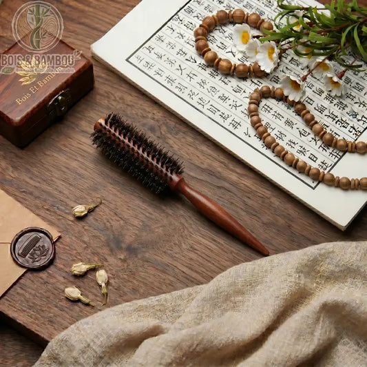 Brosse à cheveux ronde en poils de sanglier et manche en bois posée sur une table avec décor zen, calligraphie et perles. Accessoire professionnel pour brushing et volume naturel.