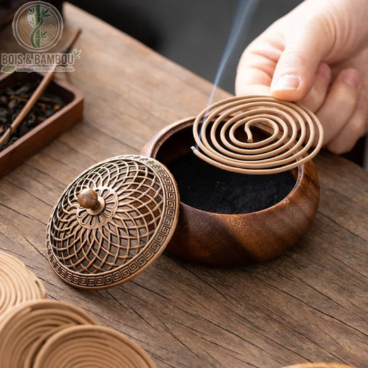 Brûleur d'Encens en Noyer Massif, un élégant récipient arrondi en bois de noyer avec un couvercle en métal finement ciselé, montré avec une main déposant une spirale d'encens allumée sur un lit de cendres noires.