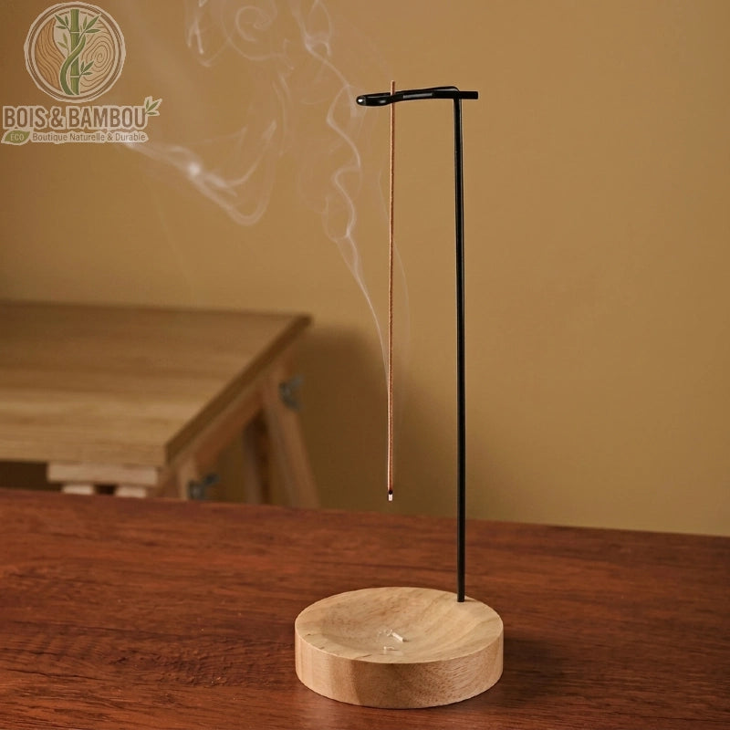 Brûleur d'encens minimaliste en bois et bambou composé d'un socle circulaire en bois clair et d'une tige métallique noire verticale. Un bâtonnet d'encens y est suspendu tête en bas, laissant s'échapper une fine volute de fumée dans une atmosphère sereine.