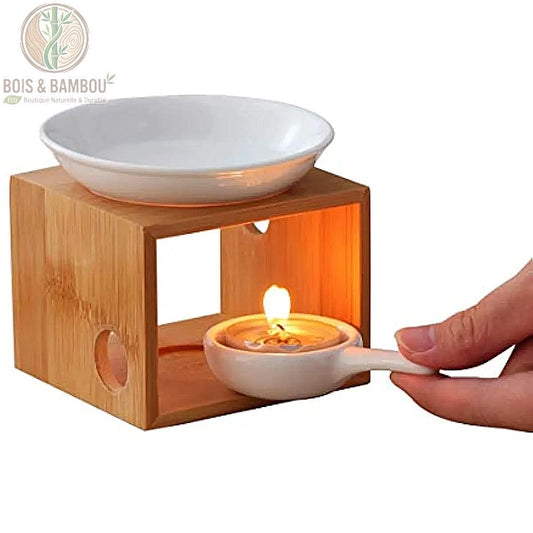 Brûleur d'huiles essentielles design avec structure cubique en bambou naturel et coupelle supérieure en céramique blanche. Une main insère un porte-bougie en céramique contenant une bougie chauffe-plat allumée à l'intérieur de la structure.