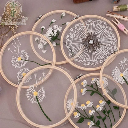 Cadre de broderie en bambou naturel mettant en valeur des motifs floraux délicats, tels que des pissenlits et des marguerites, brodés sur un voile de tulle transparent.