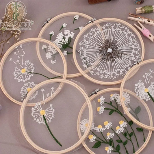 Cadre de broderie en bambou naturel mettant en valeur des motifs floraux délicats, tels que des pissenlits et des marguerites, brodés sur un voile de tulle transparent.