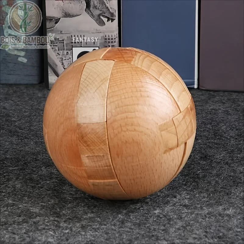 Casse-tête sphérique en bois naturel. Un puzzle complexe en forme de sphère parfaite composé de plusieurs pièces de bois imbriquées, posé sur une surface grise devant des livres.