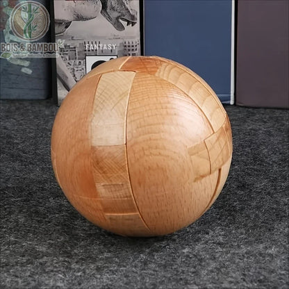 Casse-tête sphérique en bois naturel. Un puzzle complexe en forme de sphère parfaite composé de plusieurs pièces de bois imbriquées, posé sur une surface grise devant des livres.
