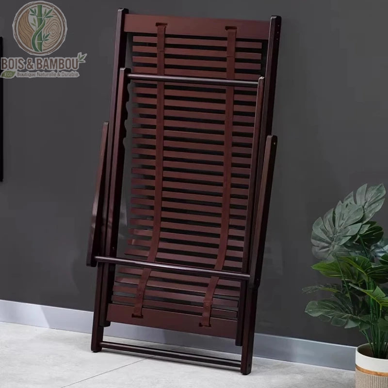 Chaise Longue en Bambou replie. Vue d'une chaise longue en bambou teinté noyer complètement repliée et appuyée contre un mur gris pour illustrer un rangement compact et pratique.