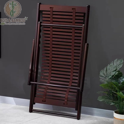 Chaise Longue en Bambou replie. Vue d'une chaise longue en bambou teinté noyer complètement repliée et appuyée contre un mur gris pour illustrer un rangement compact et pratique.
