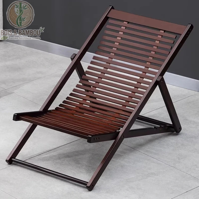 Chaise Longue en Bambou. Une chaise pliante d'extérieur en lattes de bambou de couleur brun foncé, posée sur une terrasse en dalles de pierre grise.