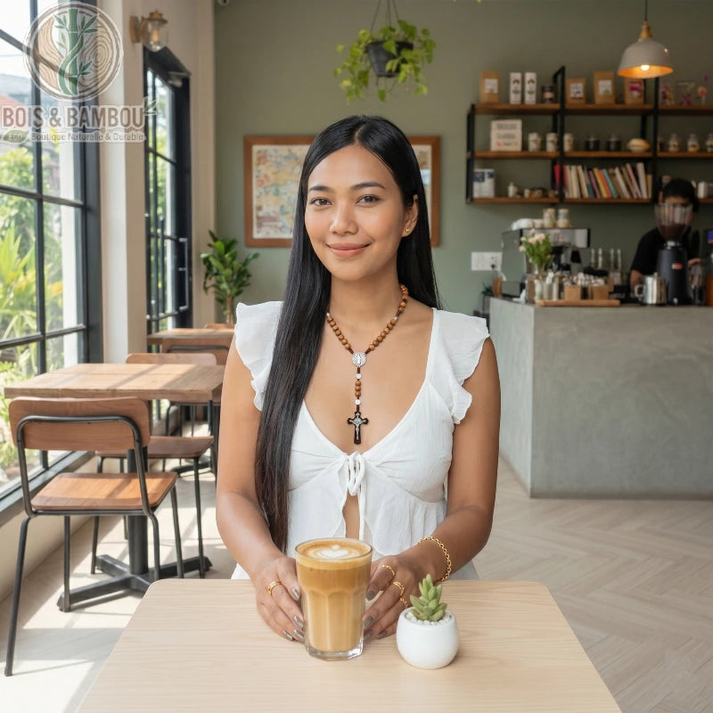 Chapelet en perles de bois naturel de 10mm avec croix, porté par une femme assise dans un café devant un latte et une petite plante.