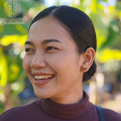 Clous d'oreilles en bois naturel et acier ornés d'un symbole Yin Yang, portés par une femme souriante de profil avec un arrière-plan naturel flou.