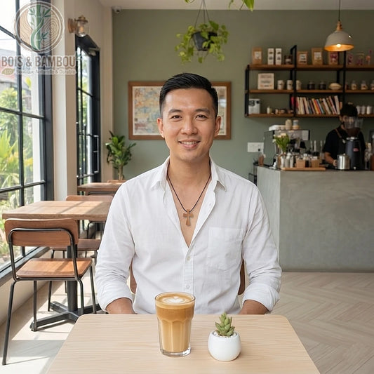 Collier croix en bois d'olivier porté par un homme souriant dans un café, assis devant un café latte et une petite plante.