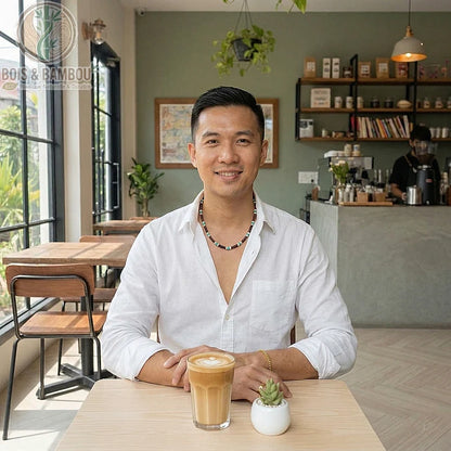 Collier de petites perles en bois porté par un homme dans un café, assis devant un café latte et une petite plante grasse.