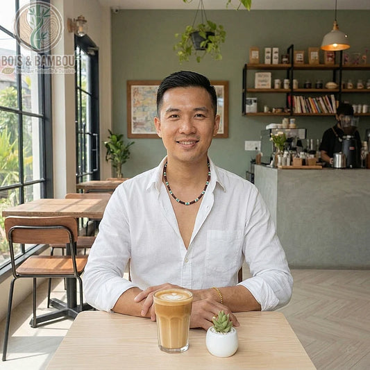 Collier de petites perles en bois porté par un homme dans un café, assis devant un café latte et une petite plante grasse.