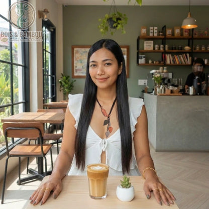 Collier avec pendentifs en forme de feuilles en bois porté par une femme dans un café, assise devant un café latte et une petite plante.
