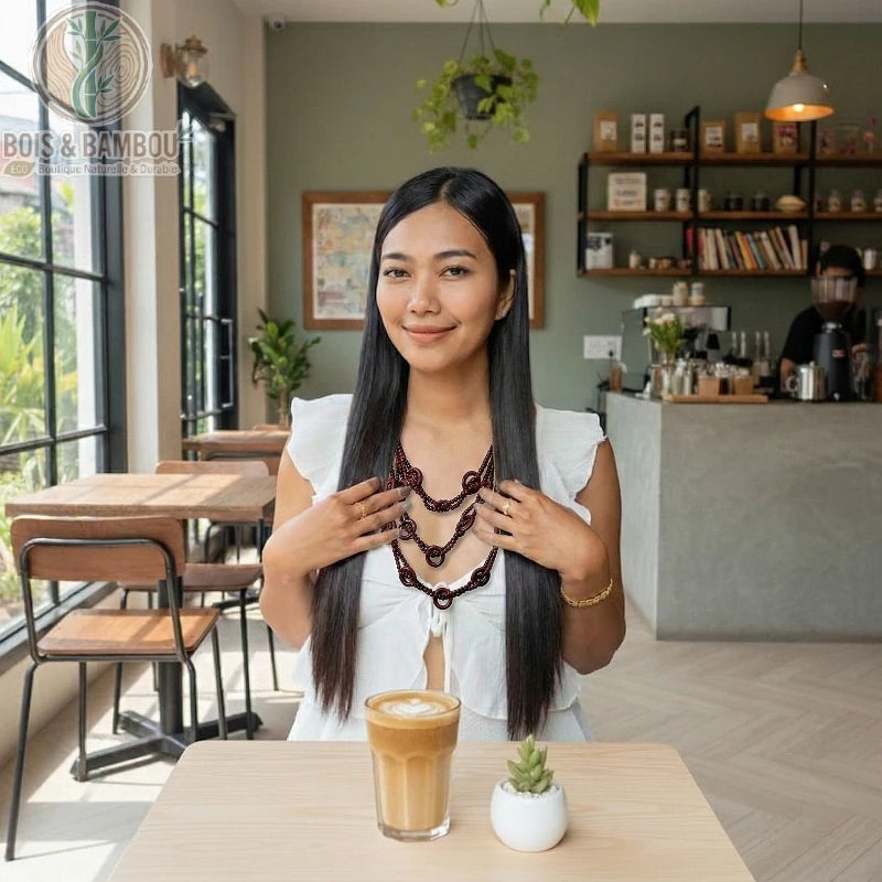 Collier multicouches en perles de bois sombre porté par une femme dans un café, assise devant un latte et une petite plante.