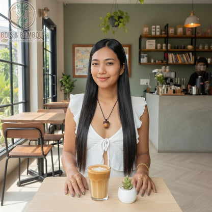 Collier avec pendentif gland en bois porté par une femme dans un café, assise devant un latte et une petite plante grasse.