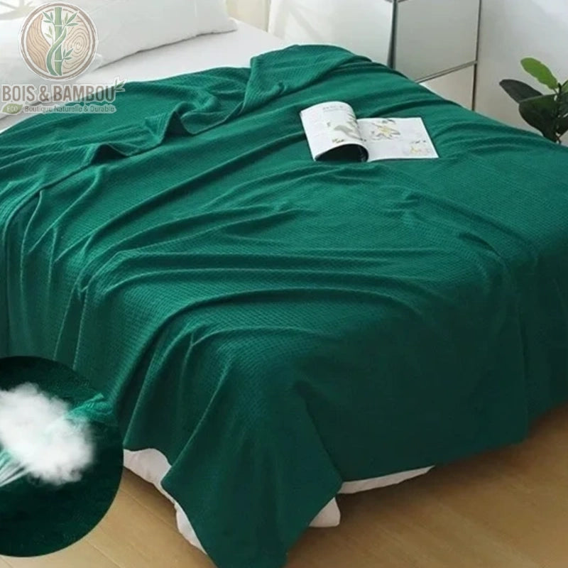 Couverture en Fibre de Bambou. Une grande couverture vert émeraude à texture gaufrée drapée sur un lit, avec un livre ouvert posé dessus. Insert montrant la douceur du rembourrage.