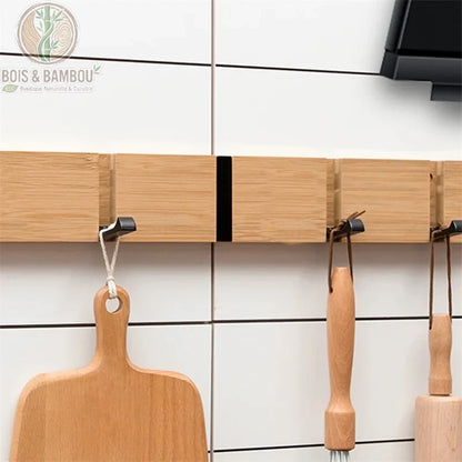 Crochets Muraux Pliables. Un support mural moderne en bambou avec des crochets noirs rabattables, utilisé pour suspendre une planche à découper et des ustensiles de cuisine.