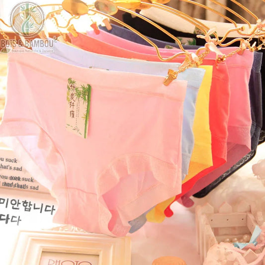 Slip bikini en laine mérinos et fibre de bambou. Plusieurs culottes aux couleurs vives (rose, bleu, jaune, corail) suspendues à des cintres dorés, affichant une bordure en dentelle.