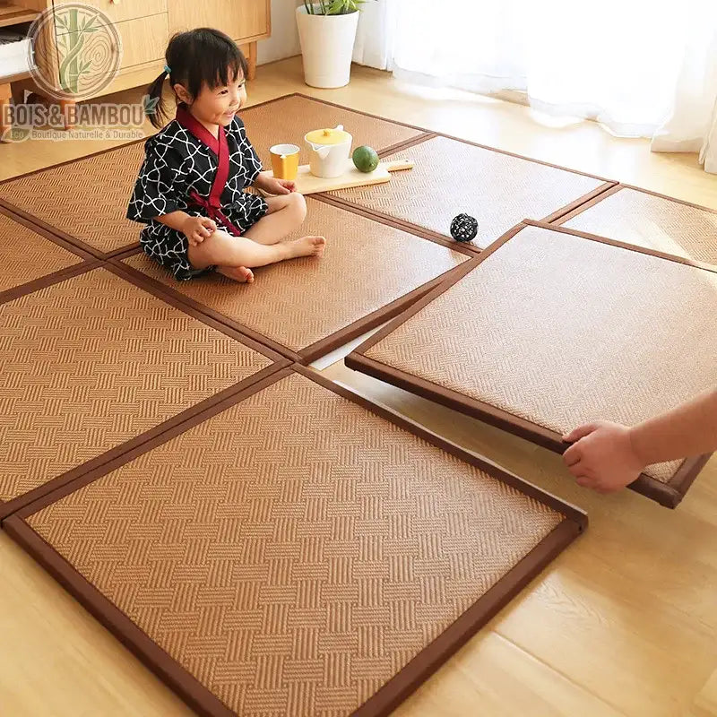 Dalles de Sol en Rotin, une petite fille assise sur un tapis modulaire composé de plusieurs dalles carrées en fibres naturelles tressées, illustrant un espace de jeu confortable et chaleureux sur un parquet en bois clair.