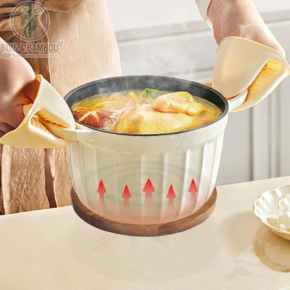 Dessous de Plat Magnétique en Acacia. Une cocotte fumante portée avec des gants en silicone est déposée sur un dessous de plat rond en bois d'acacia protégeant la surface de la chaleur.