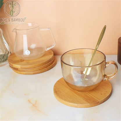 Dessous de Plat Rond en Bambou. Une tasse en verre ambré avec cuillère dorée et une carafe transparente posées sur des dessous de plat ronds en bambou clair sur un comptoir en marbre.