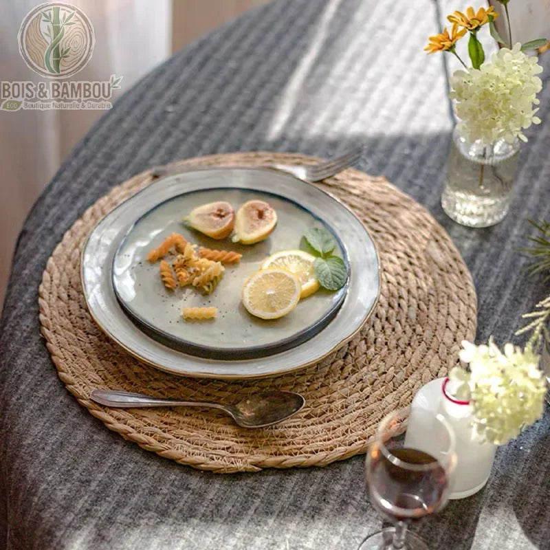Dessous de Plat en Rotin Tressé. Un set de table rond en rotin tressé supportant une assiette garnie de figues et citron sur une nappe grise. Décoration de table naturelle et bohème.