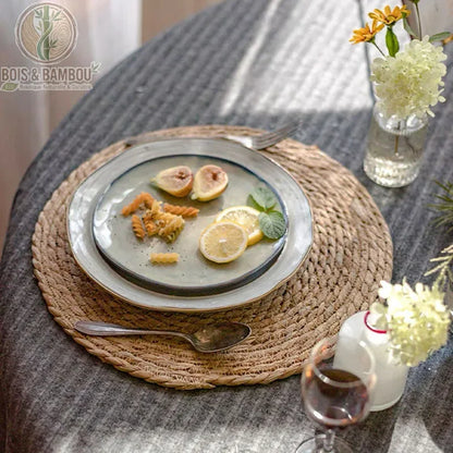 Dessous de Plat en Rotin Tressé. Un set de table rond en rotin tressé supportant une assiette garnie de figues et citron sur une nappe grise. Décoration de table naturelle et bohème.