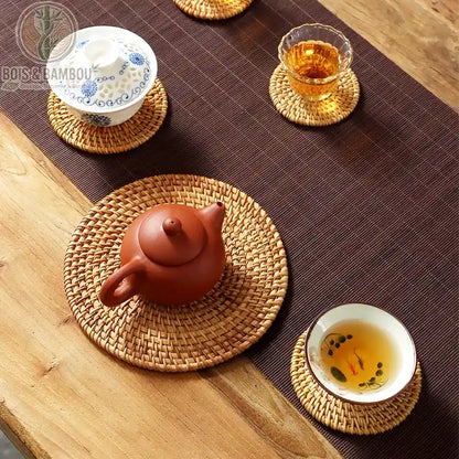 Dessous de Verre en Rotin Tissé, une scène de thé traditionnelle montrant divers sous-plats circulaires en rotin tressé à la main, supportant une théière en terre cuite, un bol à thé avec motif de poissons et une tasse en porcelaine bleue et blanche sur une table en bois.
