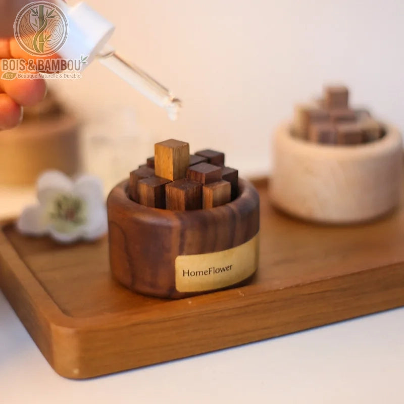 Diffuseur d'huiles essentielles en bois massif, un socle circulaire en bois sombre portant l'inscription "HomeFlower", rempli de petits cubes de bois de différentes teintes, présenté avec une pipette versant une goutte d'huile essentielle pour une diffusion passive sur un plateau en bois.