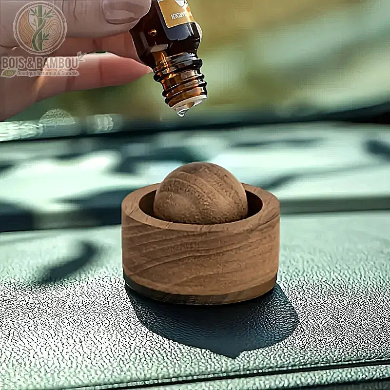 Diffuseur d'huiles essentielles en bois naturel, un petit socle circulaire en bois clair contenant une bille de bois assortie, illustré avec une main versant une goutte d'huile essentielle directement sur la bille pour une diffusion naturelle dans l'habitacle d'une voiture.