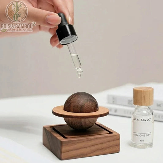 Diffuseur de parfum en bois sculpté Jupiter, un diffuseur passif original composé d'une sphère et d'un anneau en bois évoquant une planète, reposant sur un socle carré, illustré avec une main utilisant une pipette pour déposer de l'huile essentielle.