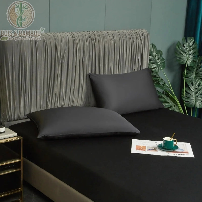 Drap-Housse en Satin de Bambou Noir 160x200 cm. Lit double élégant paré d'un drap-housse noir profond et d'oreillers assortis au fini satiné, devant une tête de lit plissée grise.