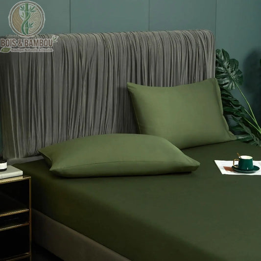 Drap-Housse en Satin de Bambou Vert 180x200 cm. Un lit double paré d'un drap-housse et d'oreillers vert forêt au fini satiné, devant une tête de lit plissée grise et un mur sombre.