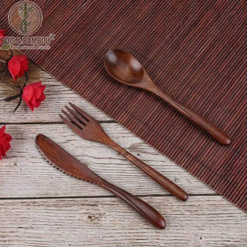 Ensemble de vaisselle en bois réutilisable. Un set comprenant une cuillère, une fourchette et un couteau dentelé en bois sombre, disposés sur une table rustique avec un set en bambou.