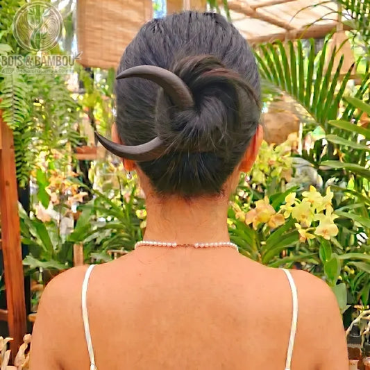 Épingle à Cheveux Croissant de Lune en bois sombre fixant le chignon d'une femme de dos, avec un collier de perles et un fond de jardin tropical.